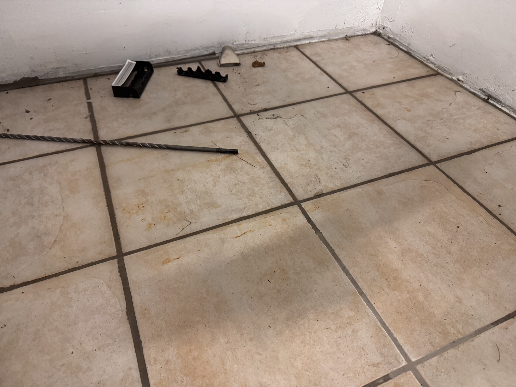 Tile floor corner with tools: long drill rod, black holder, metal wedge, orange stains.
