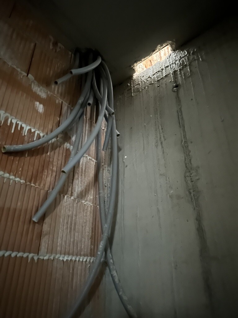 Construction site corner: gray cable conduits hanging in front of raw masonry on the left and concrete wall on the right.