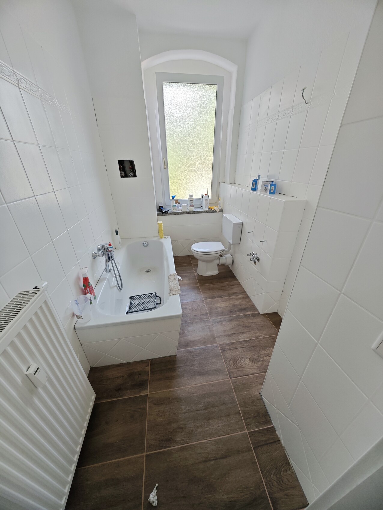 Bright bathroom with white tiled wall, bathtub on the left, window at the back, toilet, brown floor.