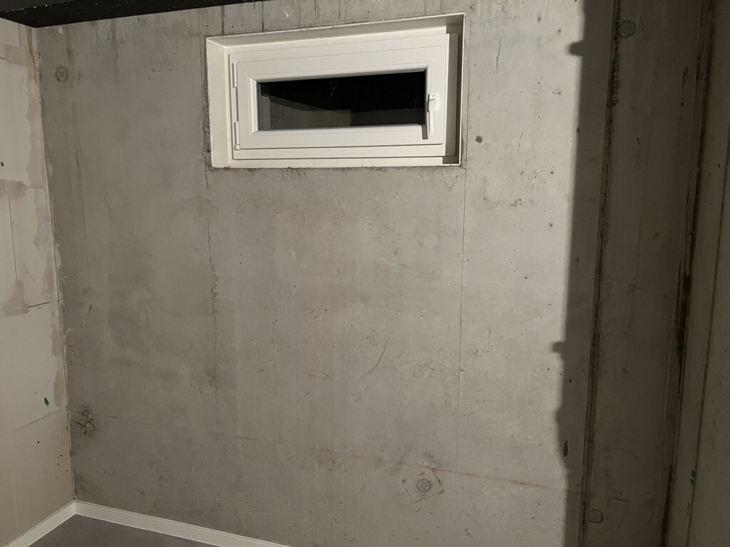 Small white horizontal window in raw concrete basement wall.