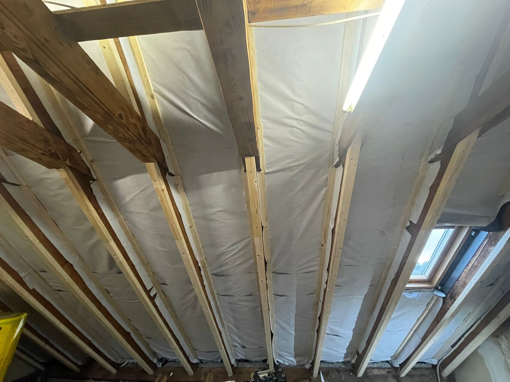 Attic with exposed wooden beams, insulation installed between them, window on the right, and fluorescent light.