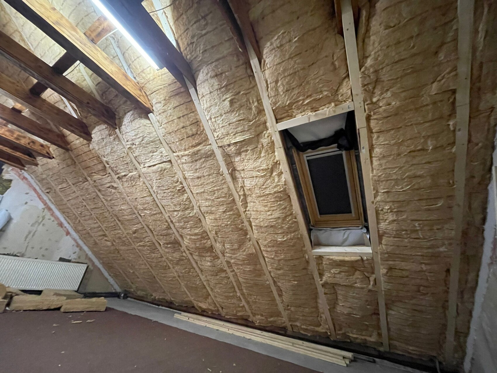 Attic with insulation between rafters, roof window and construction wood on the floor.