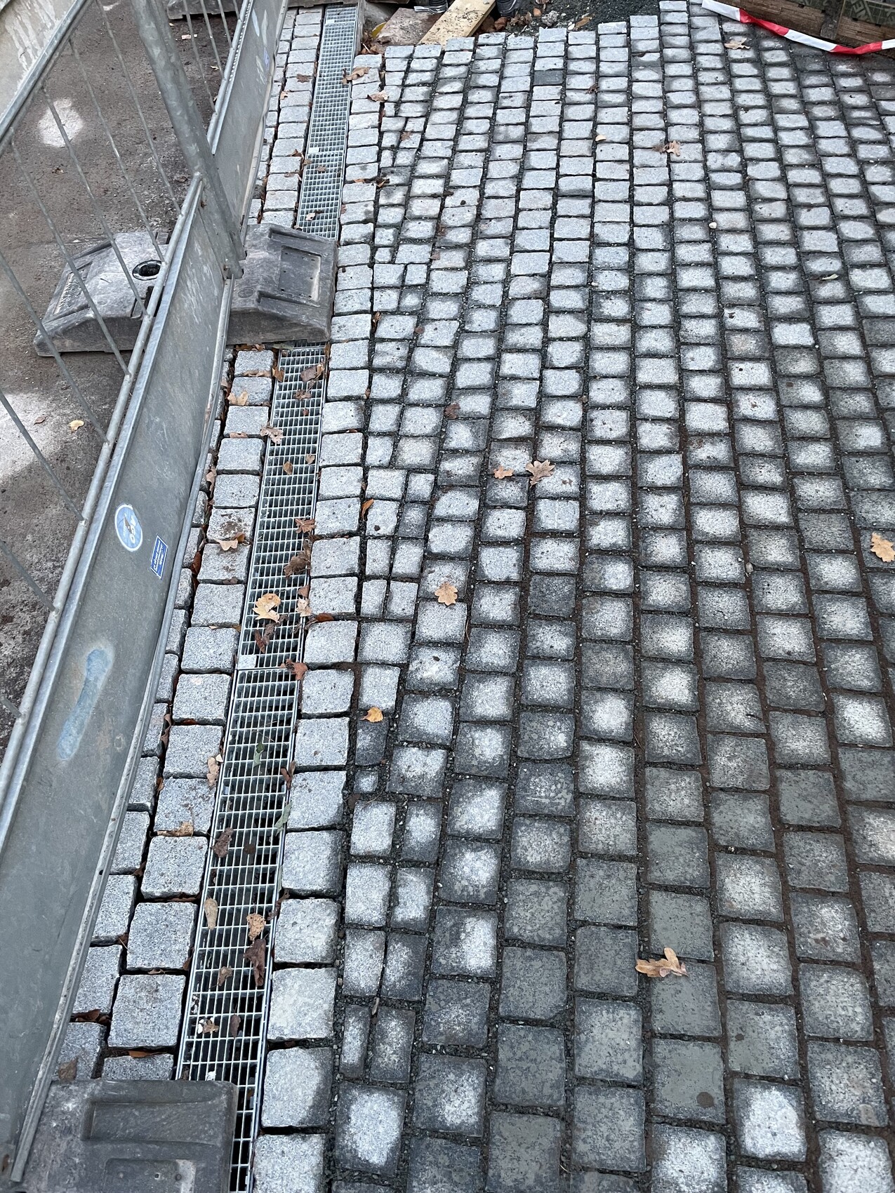 Gray sidewalk made of cobblestones with metal drainage channel at the edge, construction fence on the left, autumn leaves.