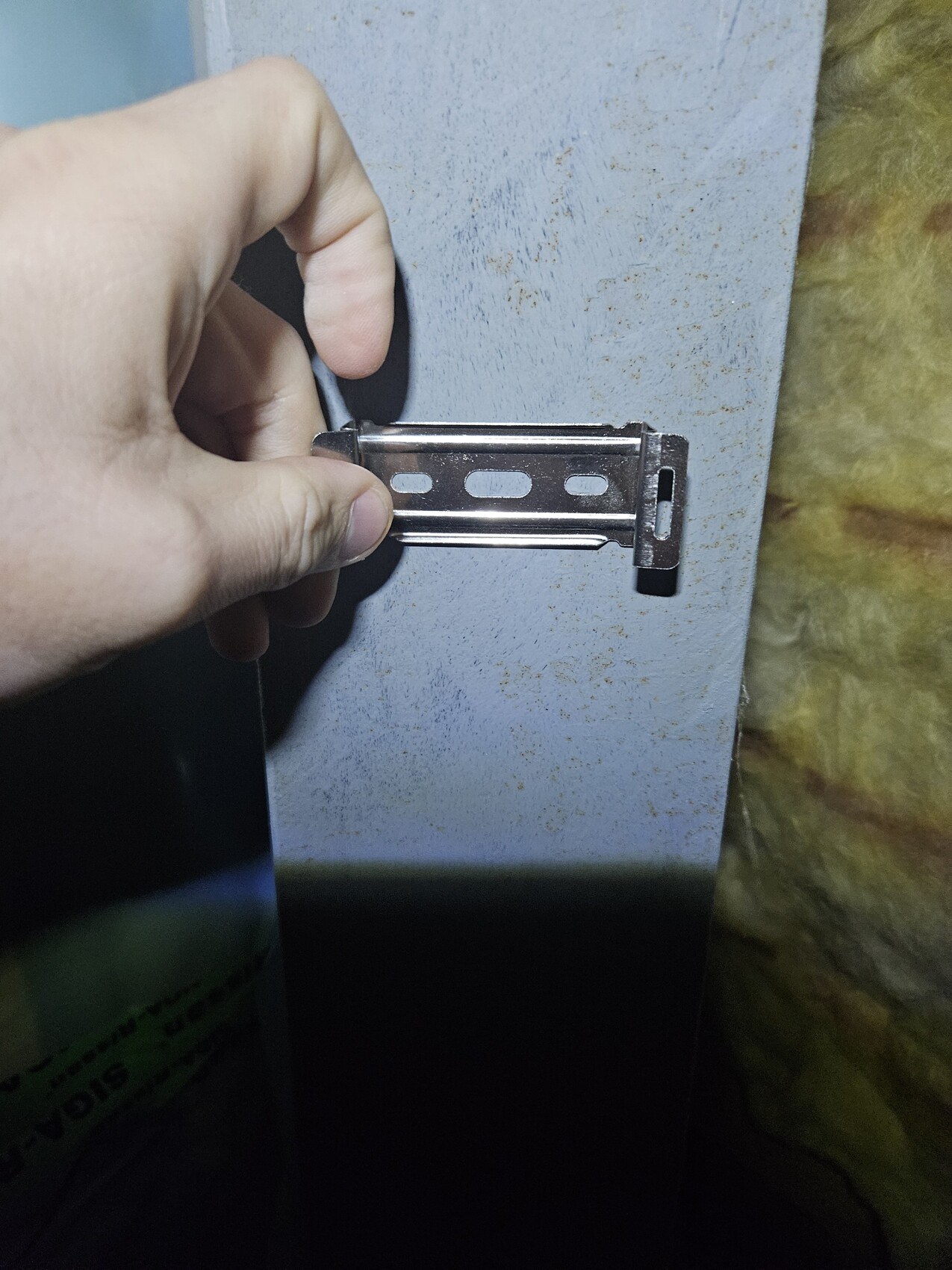 Hand holding a flat metal bracket with three oval holes in front of a bluish-gray metal surface