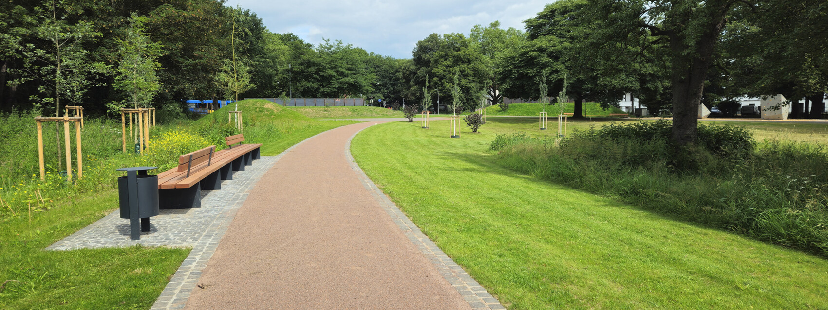 Curved park path with wooden benches on the left, trash bin, grass areas and trees.