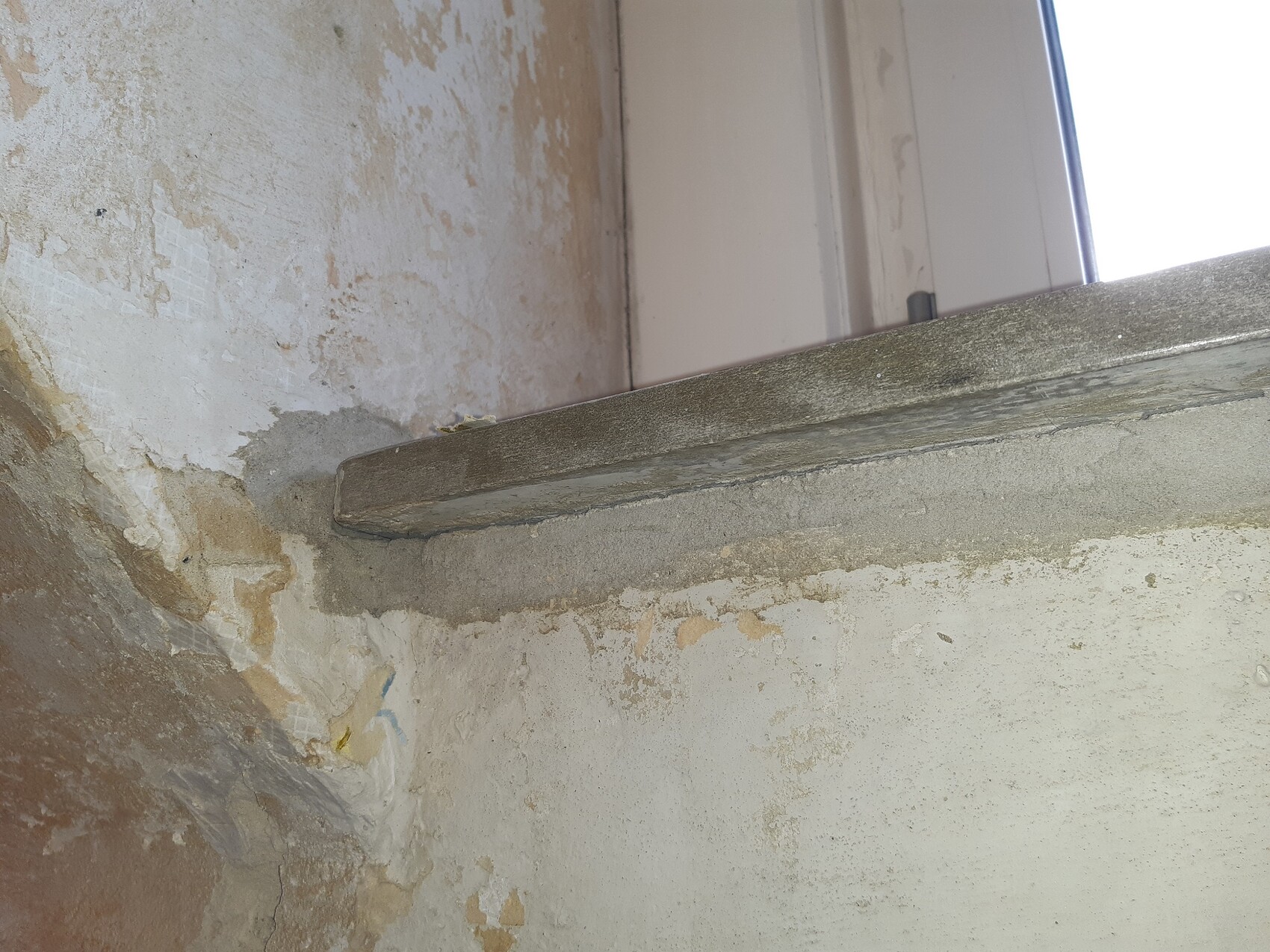 Rough wall section with peeling plaster; a gray concrete ledge protrudes as a windowsill.