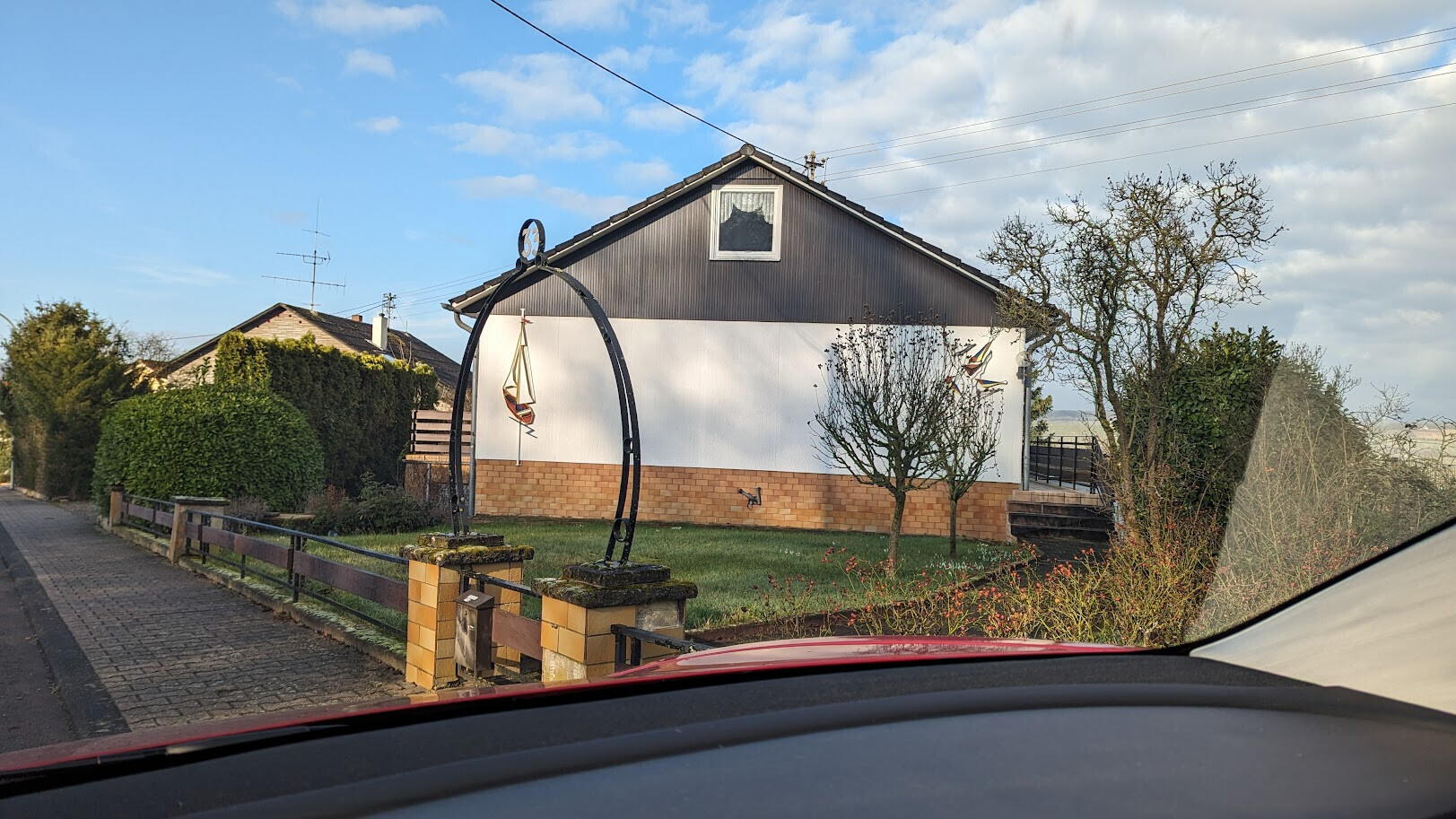 Residential house with white walls, brown base, garden and metal arch with sailboat model in the front garden.