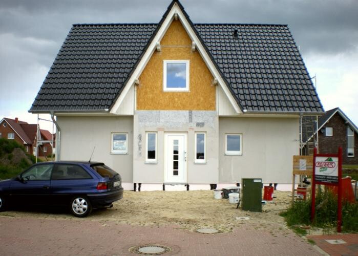 Two-story house under construction with yellow facade, dark tiled roof, car on the left.