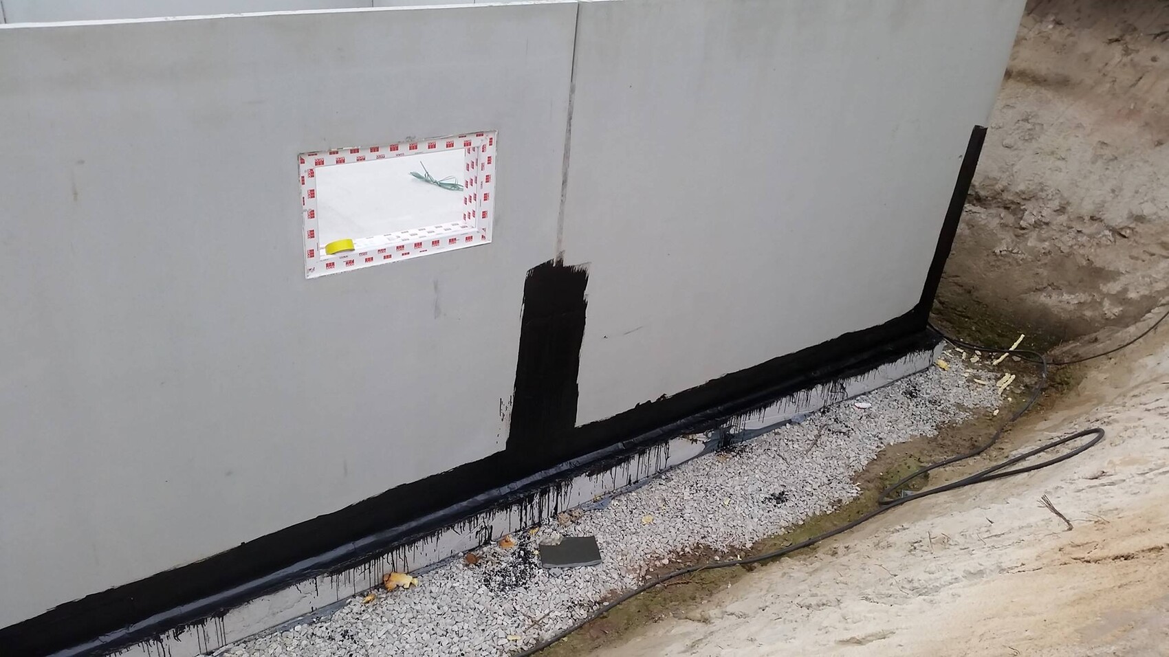 Exterior wall with black foundation waterproofing at the base, window opening, and gravel floor