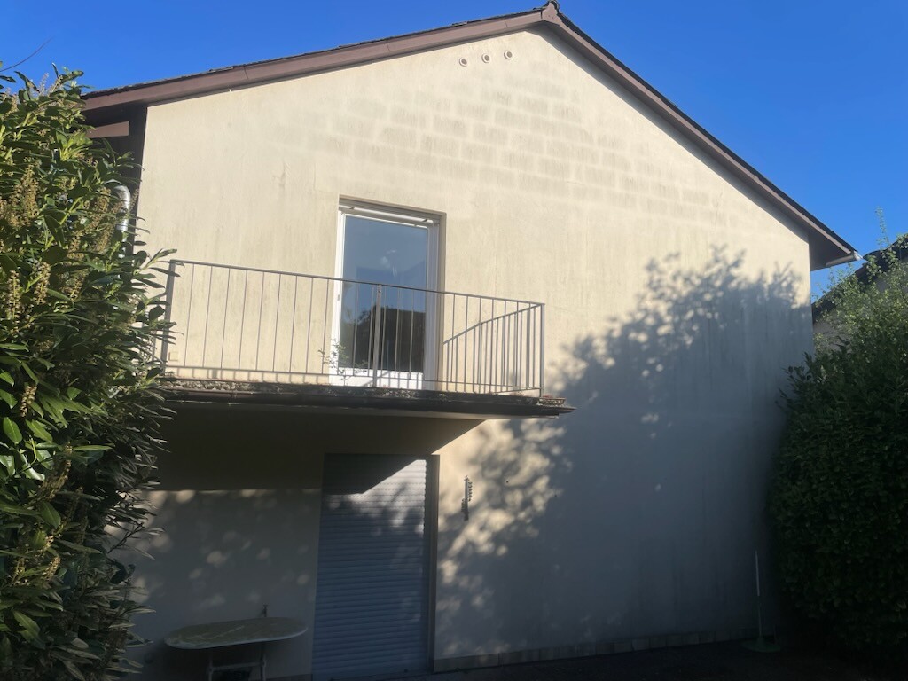 Two-story house with balcony, glass door and metal railing, surrounded by hedges