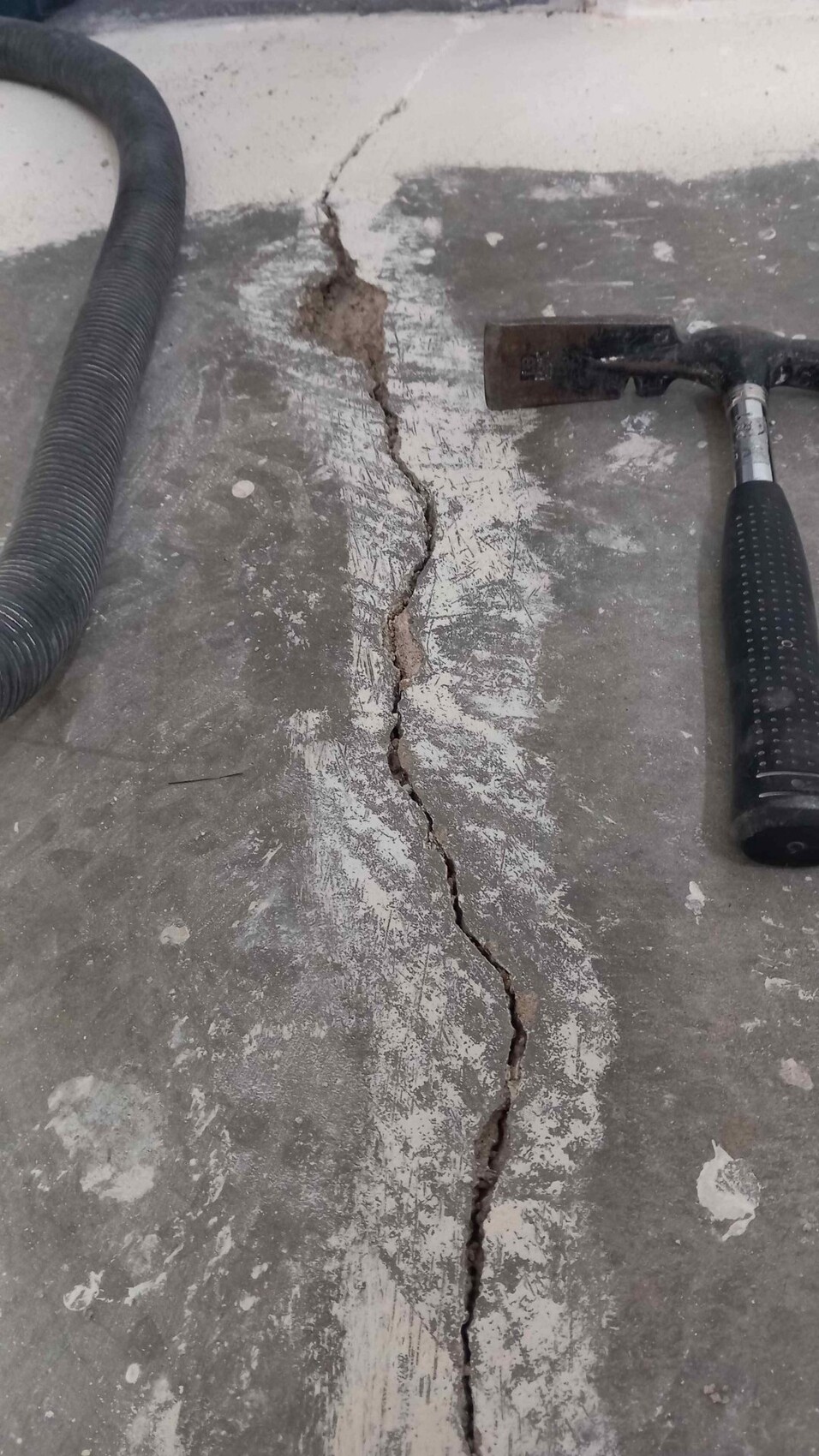 Wide crack in a concrete floor slab, hose and hammer lying next to it.