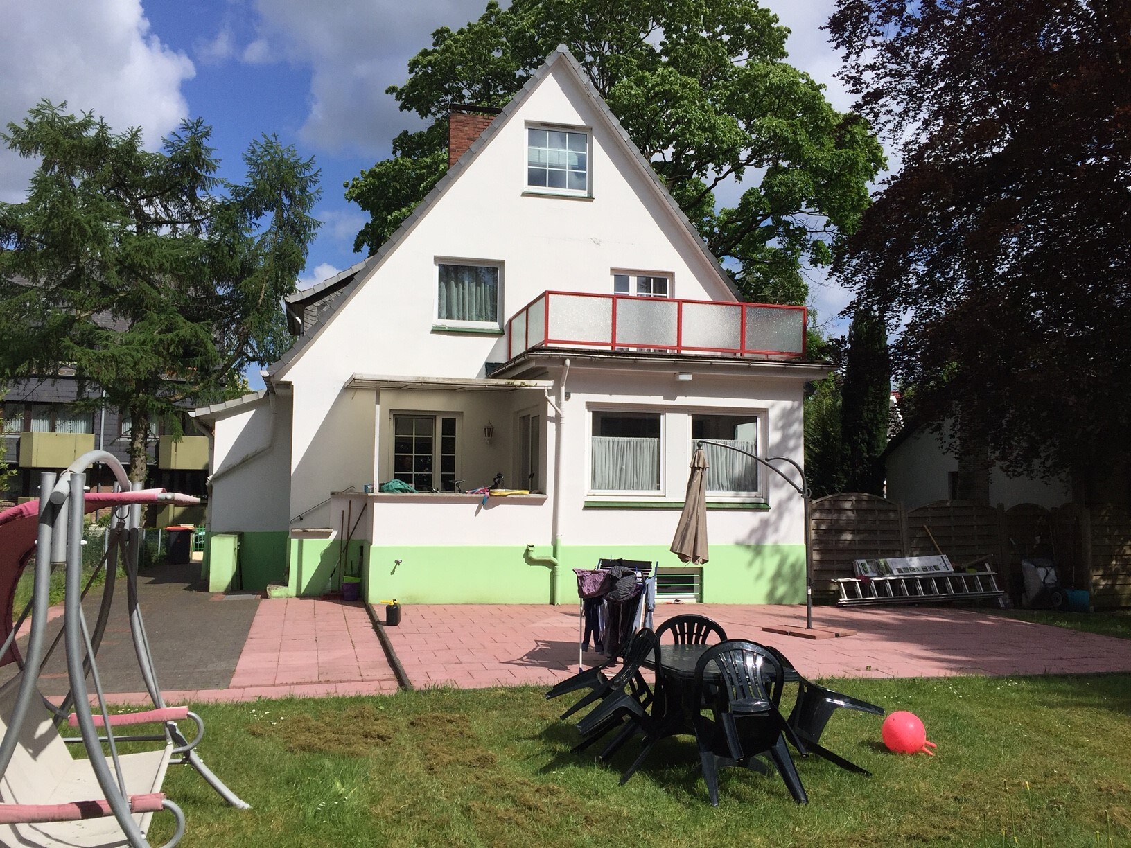 Weißes, zweistöckiges Haus mit grünem Sockel und roter Balkonbrüstung; Garten mit Stühlen.