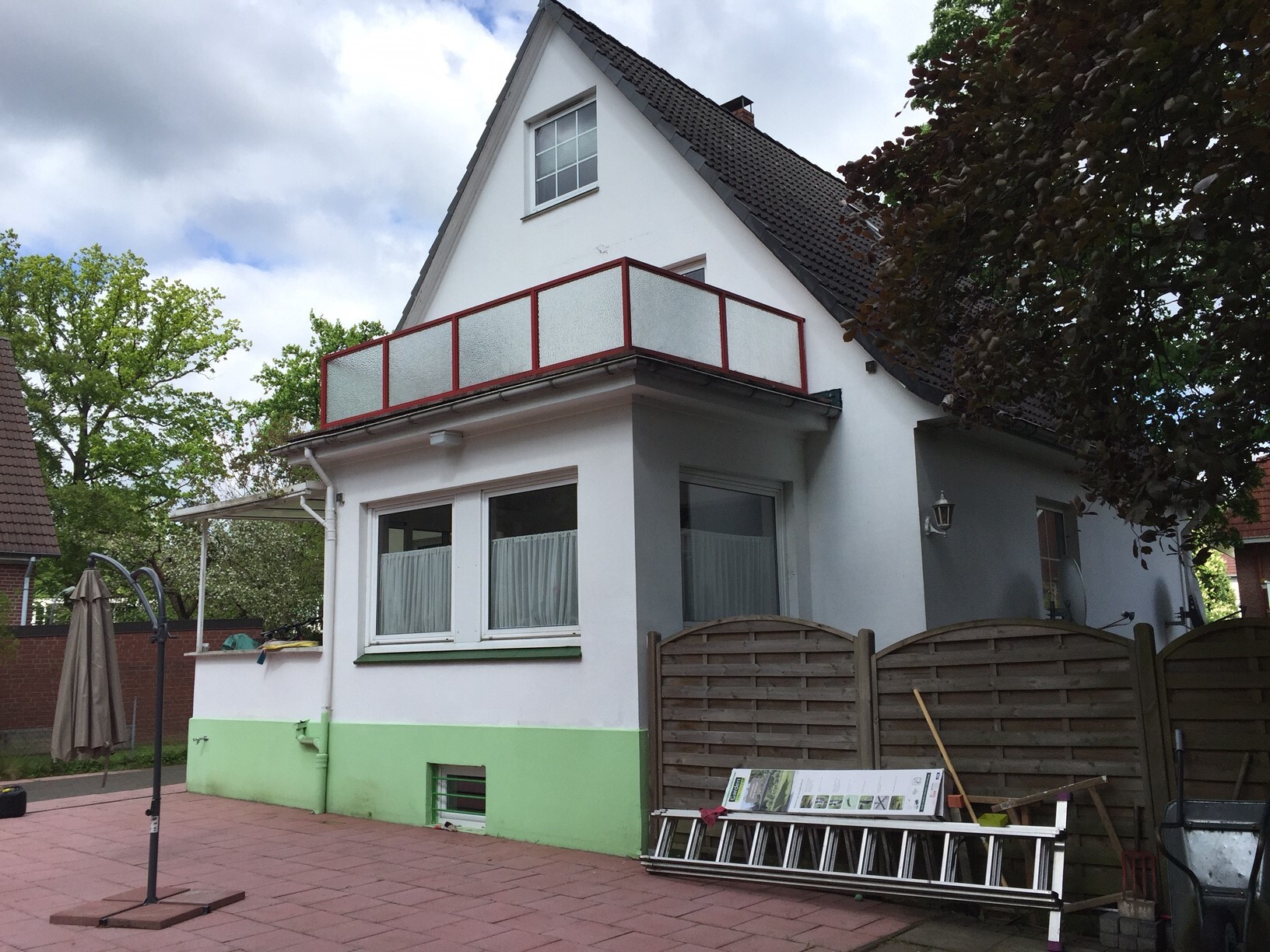 Weißes Haus mit grünem Sockel, rotem Balkongeländer, Gartenbereich, Leiter und Baumaterialien.