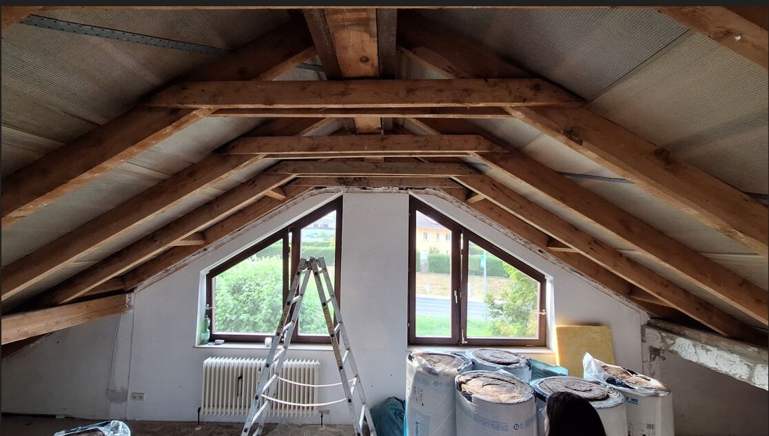 Attic renovation: visible roof beams, ladder, triangular windows, insulation material.