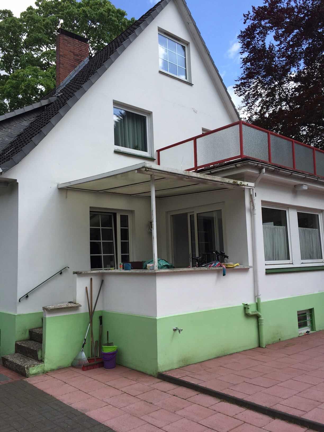 Weiße Hausfront mit grünem Sockel, Balkon mit rotem Geländer aus Glas, Gartengeräten vor Eingang