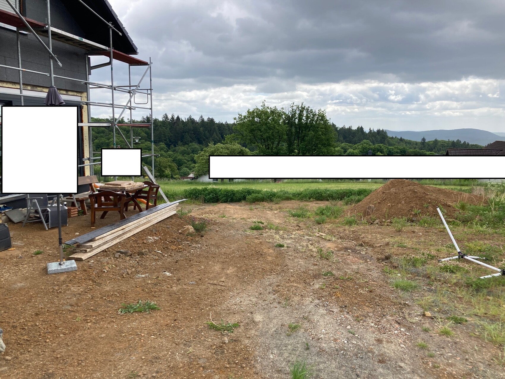 Baugerüst am Haus; Baustelle mit Holzplanken, Tisch und Stühlen, Landschaft im Hintergrund.