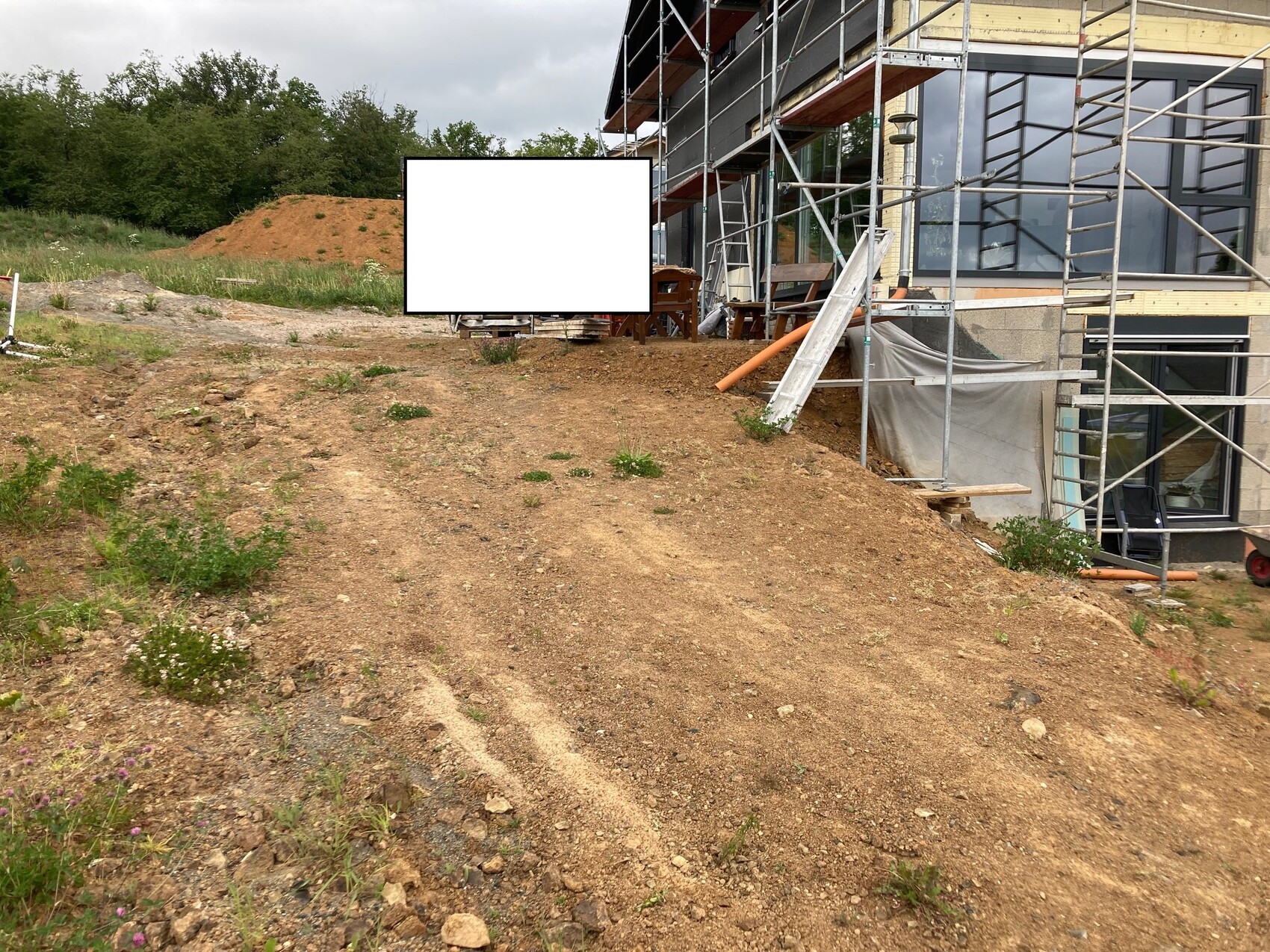 Baustelle mit Gerüst vor modernem Gebäude; unbefestigte Erdfläche und leere Werbetafel