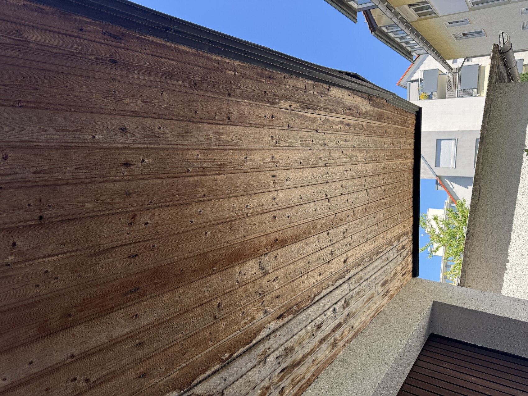 Underside of a wooden roof overhang with wooden battens; blue sky and neighboring building in the background.