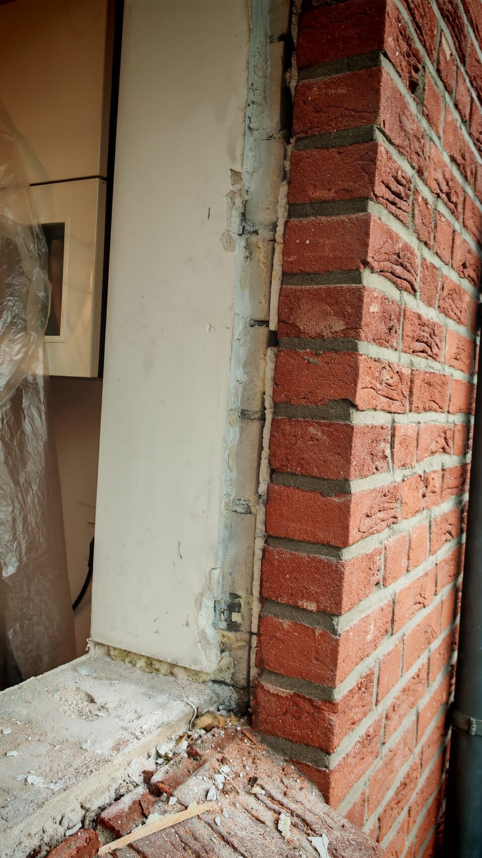 Rote Backsteinwand mit beschädigtem Putz und herumliegendem Bauabfall neben einer Türöffnung.