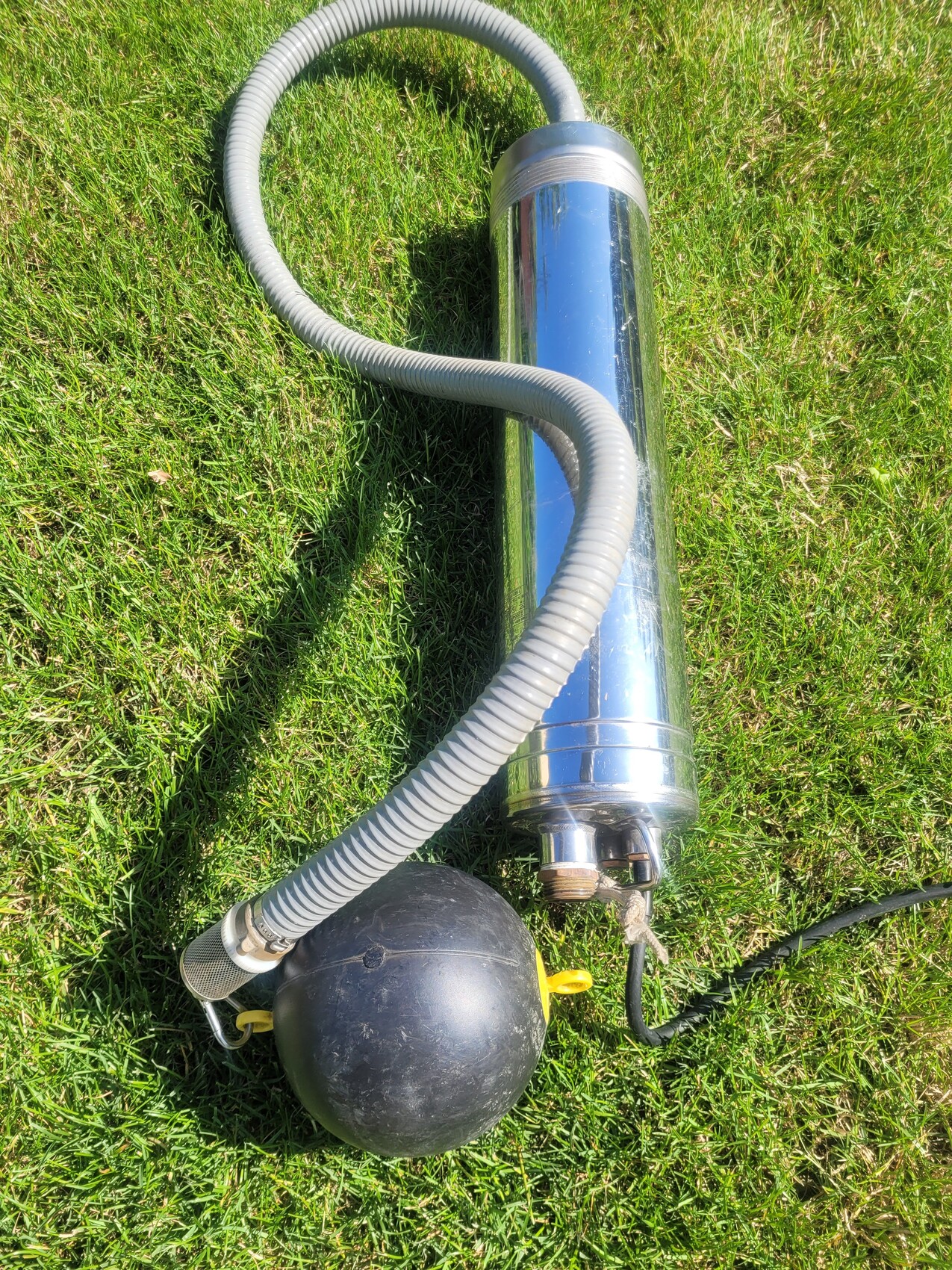 Submersible pump made of stainless steel with grey spiral hose on grass; black float ball next to it.