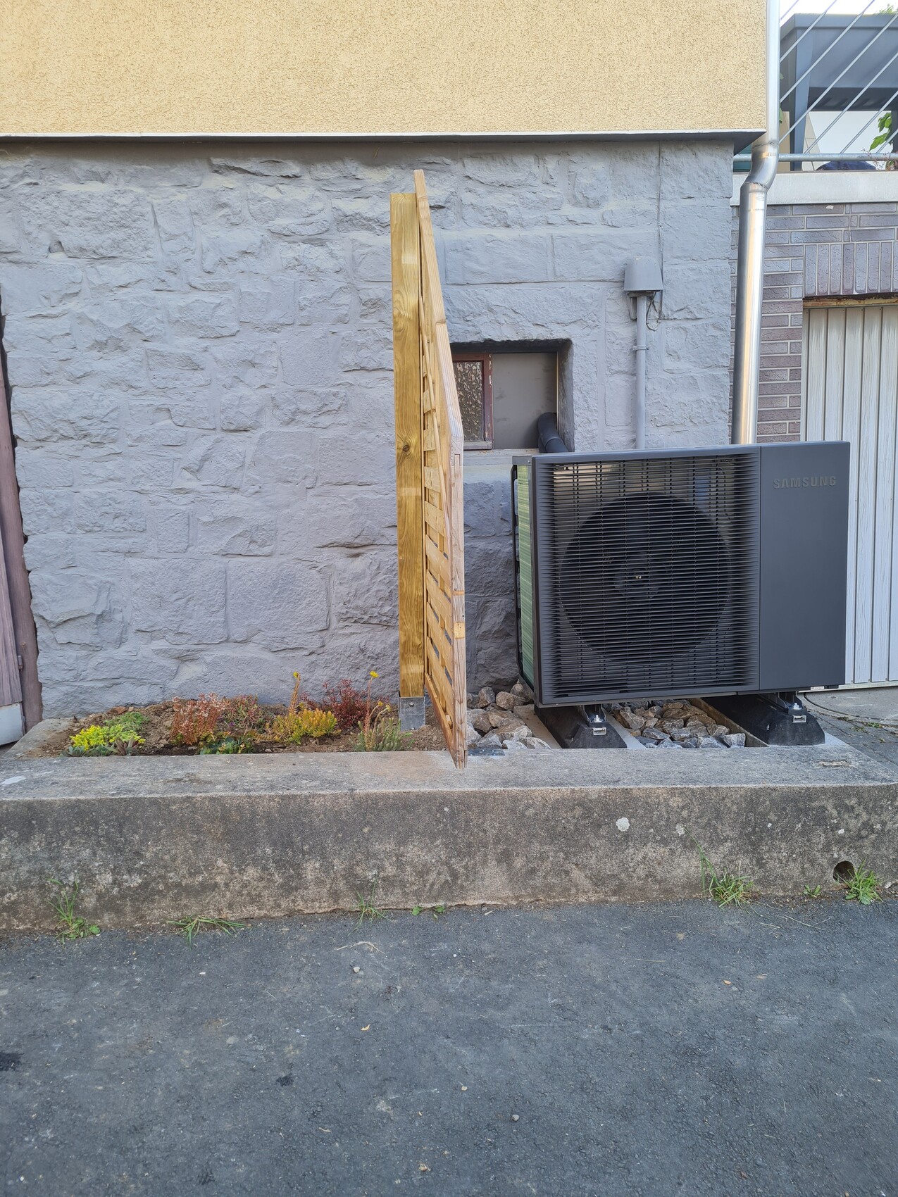 Outdoor Samsung air conditioning unit in front of gray stone wall; slanted wooden panel next to it, bed with plants.