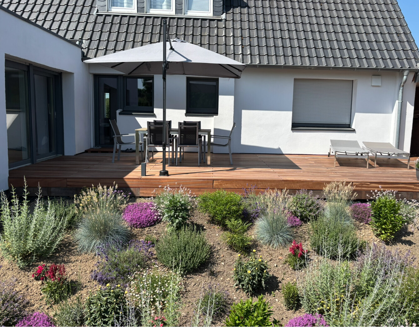 Wood deck terrace of a house with dining table under a sun umbrella, two lounge chairs, and blooming flower bed.
