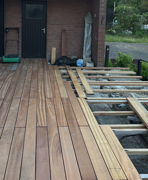 Wooden terrace being installed in front of a brick house; decking boards lie on the substructure.
