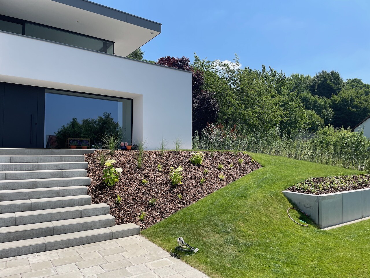 Modern white villa with dark front door, large windows, steps on the left, and a hillside garden.
