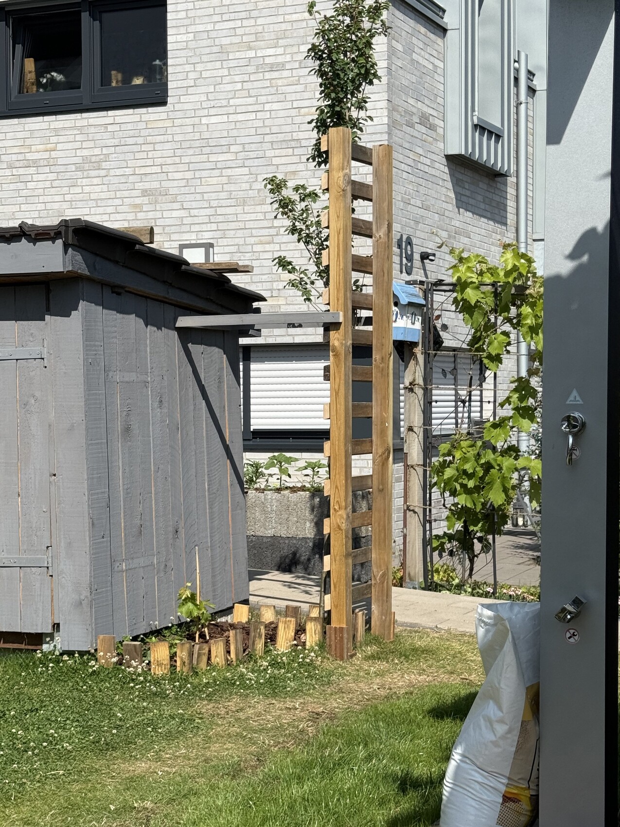 Gartenbereich neben hellem Ziegelhaus: Holzpfosten-Gitter, Kletterpflanze, graues Gartenhaus und Gras.