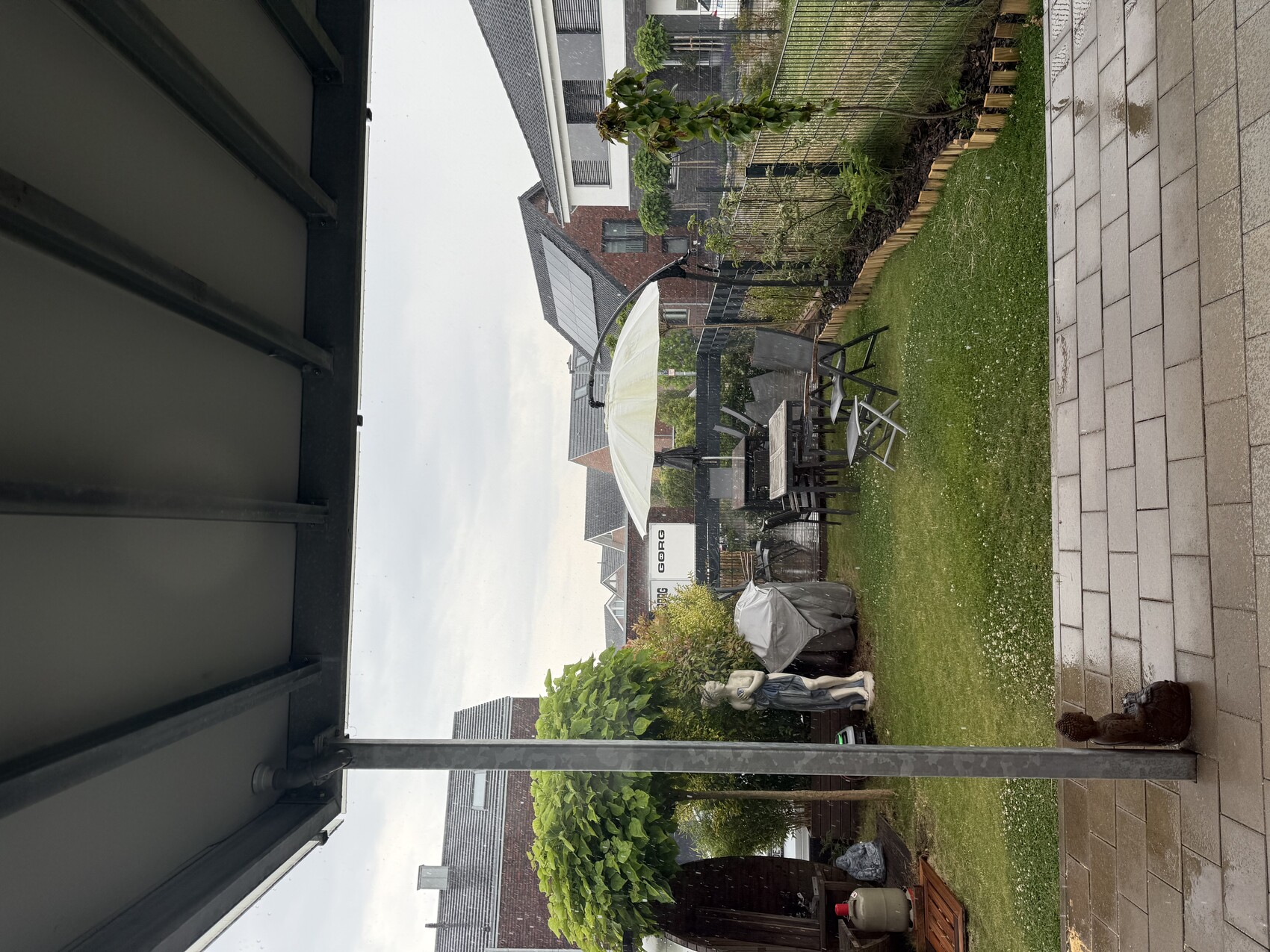 Garten hinter Terrasse: Esstisch mit Stühlen unter weißem Schirm, Grillabdeckung, Statue, Regen.