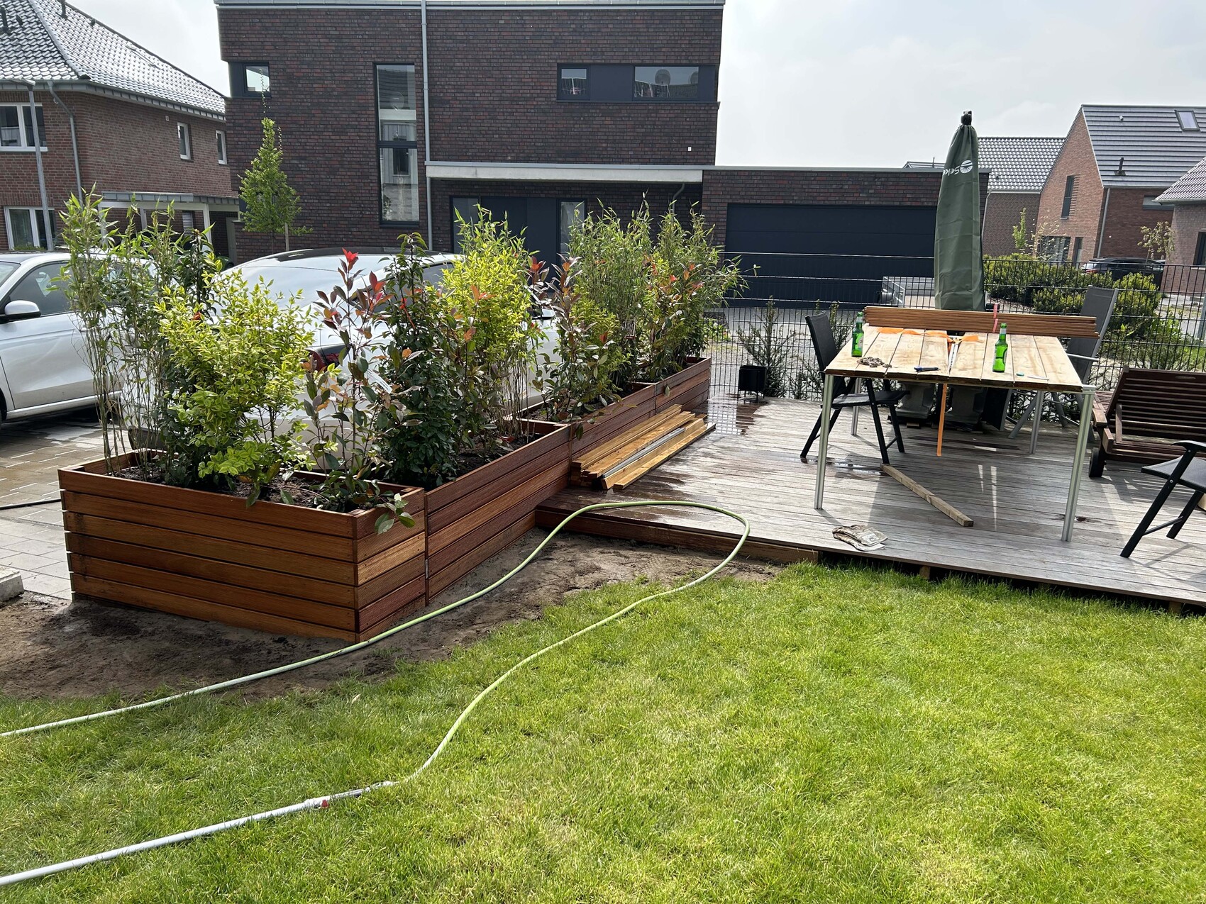 Garten mit Holzkasten voller Sträucher, Holzdeck mit Esstisch und geschlossenem Sonnenschirm.