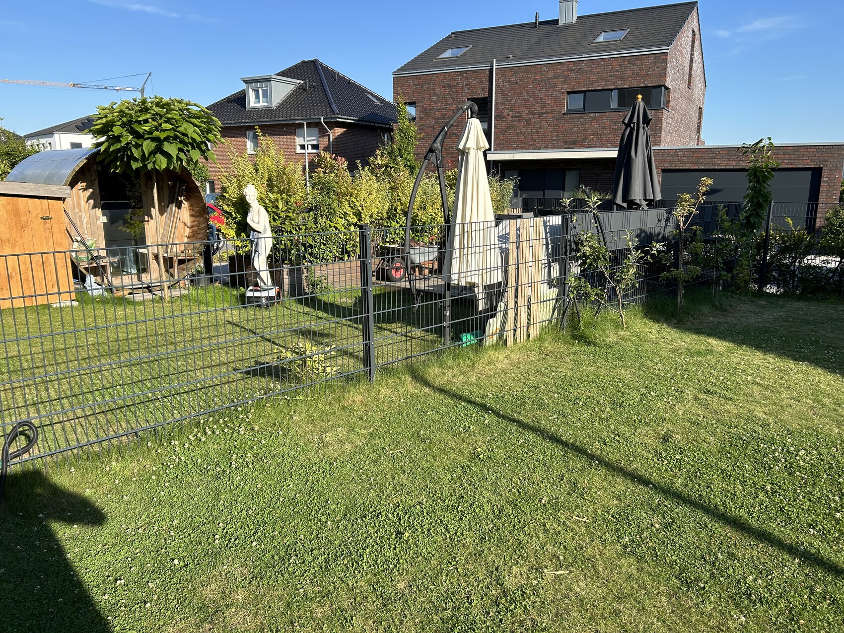 Garten hinter Metallzaun mit Sitzbereich, Statue und zwei Sonnenschirmen, Häuser im Hintergrund.