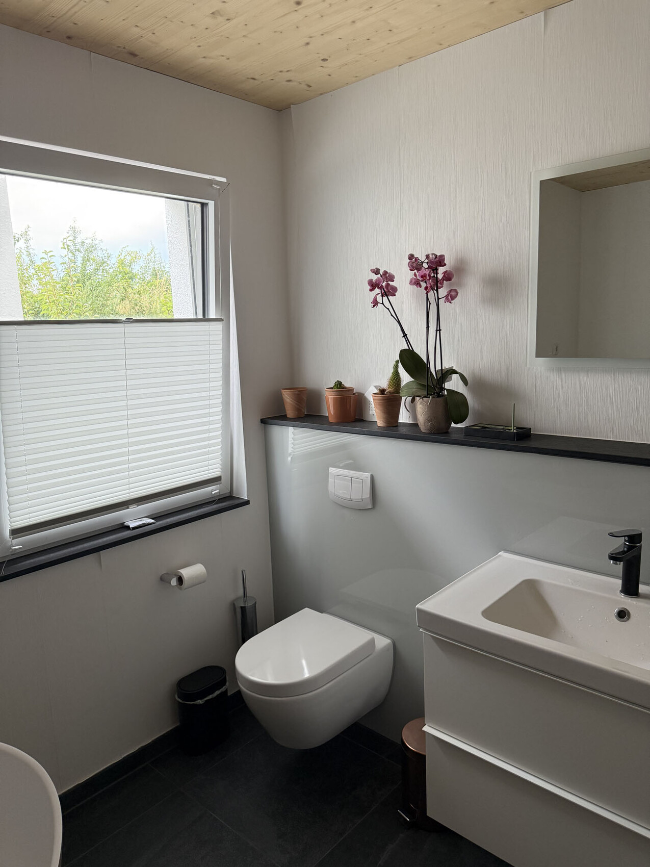 Bright bathroom with window, wall-mounted toilet, vanity, orchid on shelf, mirror.