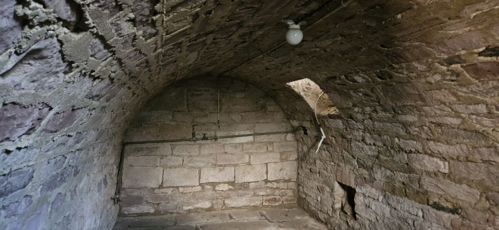 Dark stone vaulted cellar with arch, visible pipes, light bulb on the ceiling, opening on the right wall