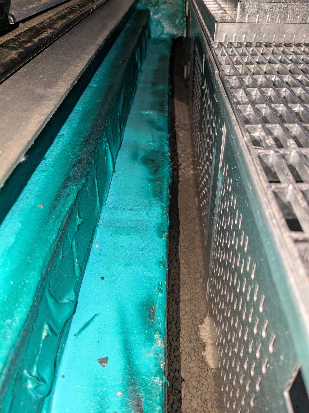 Narrow channel with turquoise side wall on the left, metal grate on the right, and gravel floor in between.