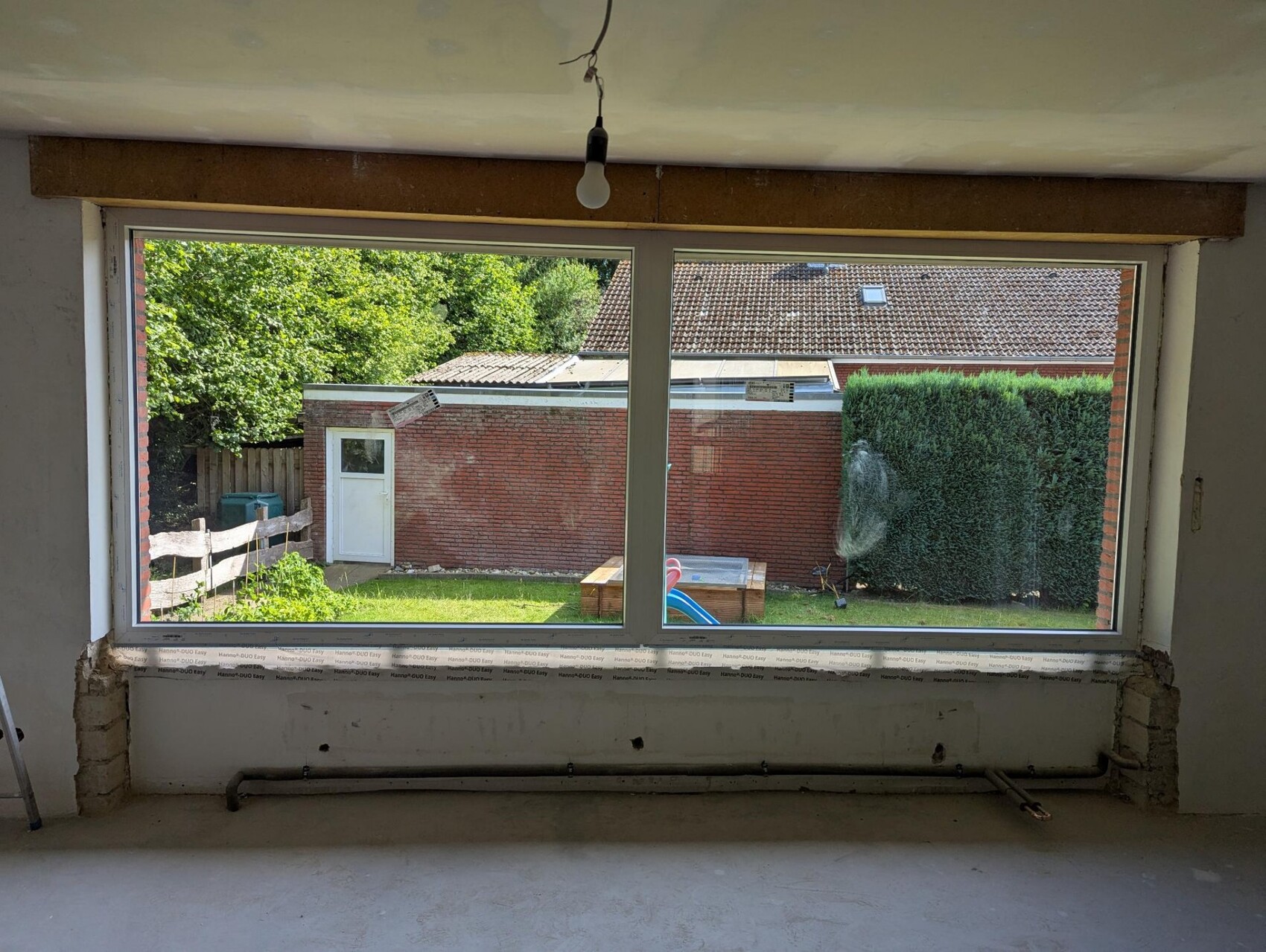 Interior of a construction site with a large window front, view of a garden with hedge, playground equipment.