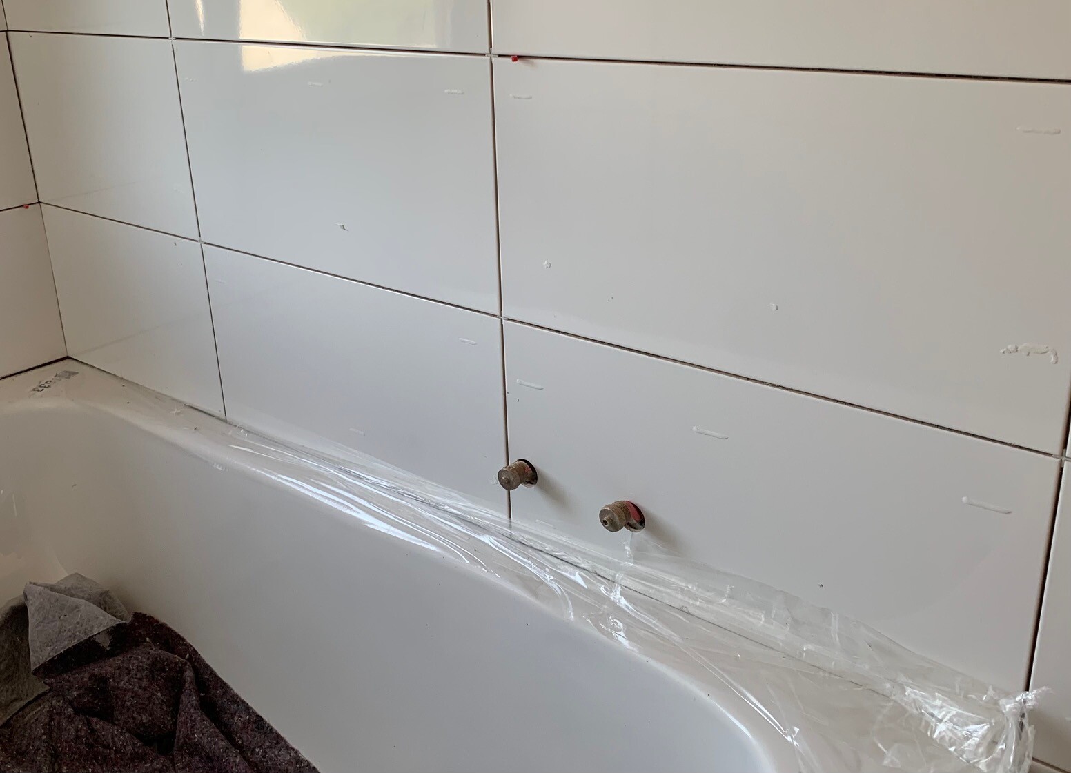 Badezimmer mit weißen Fliesen, zwei Wasseranschlüsse über der Badewanne, Schutzfolie am Rand.