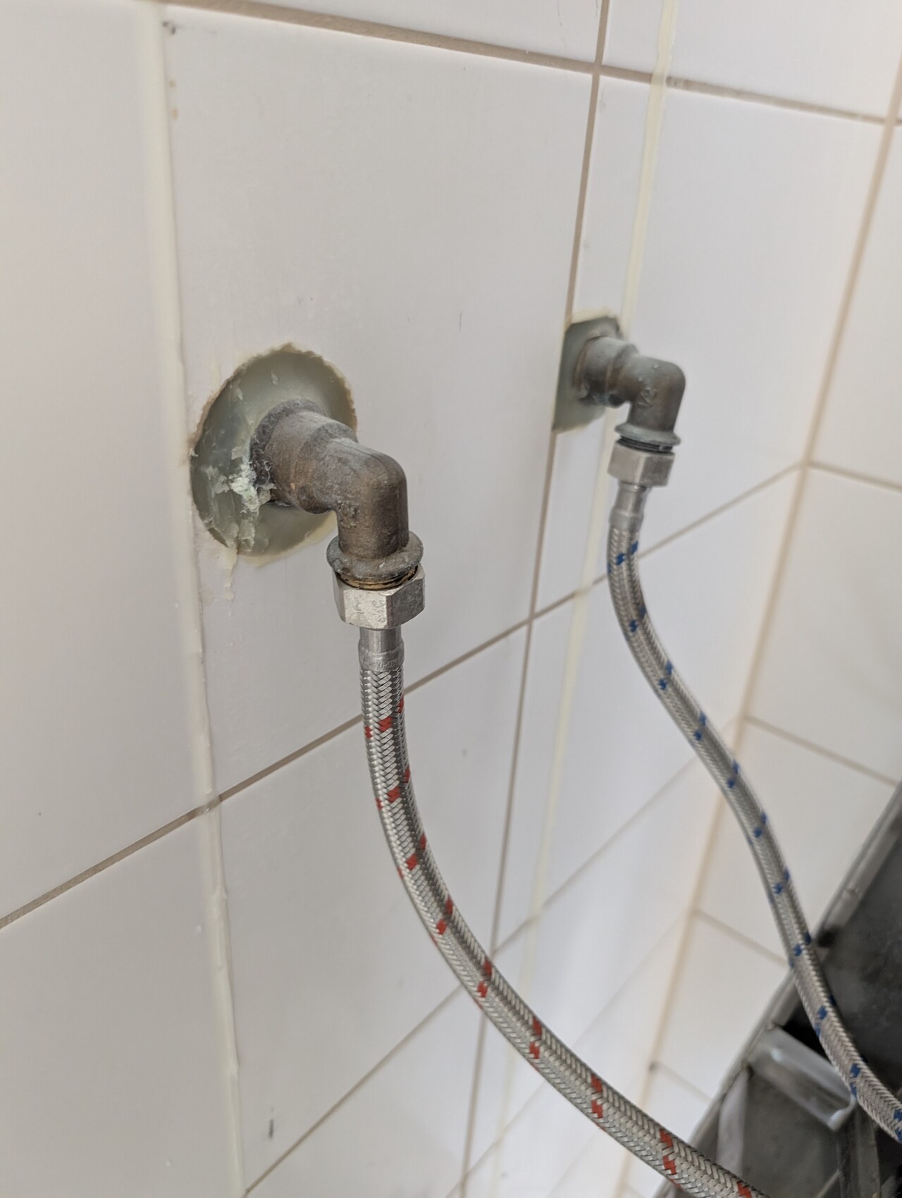 Two rusty water connections with flexible stainless steel hoses behind tiles, shut-off valves.