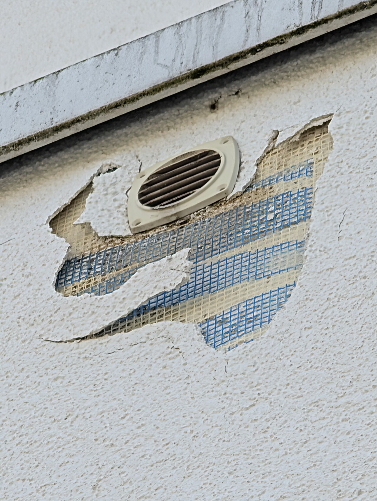 Exterior wall with peeling plaster, behind it blue mesh and round ventilation slot