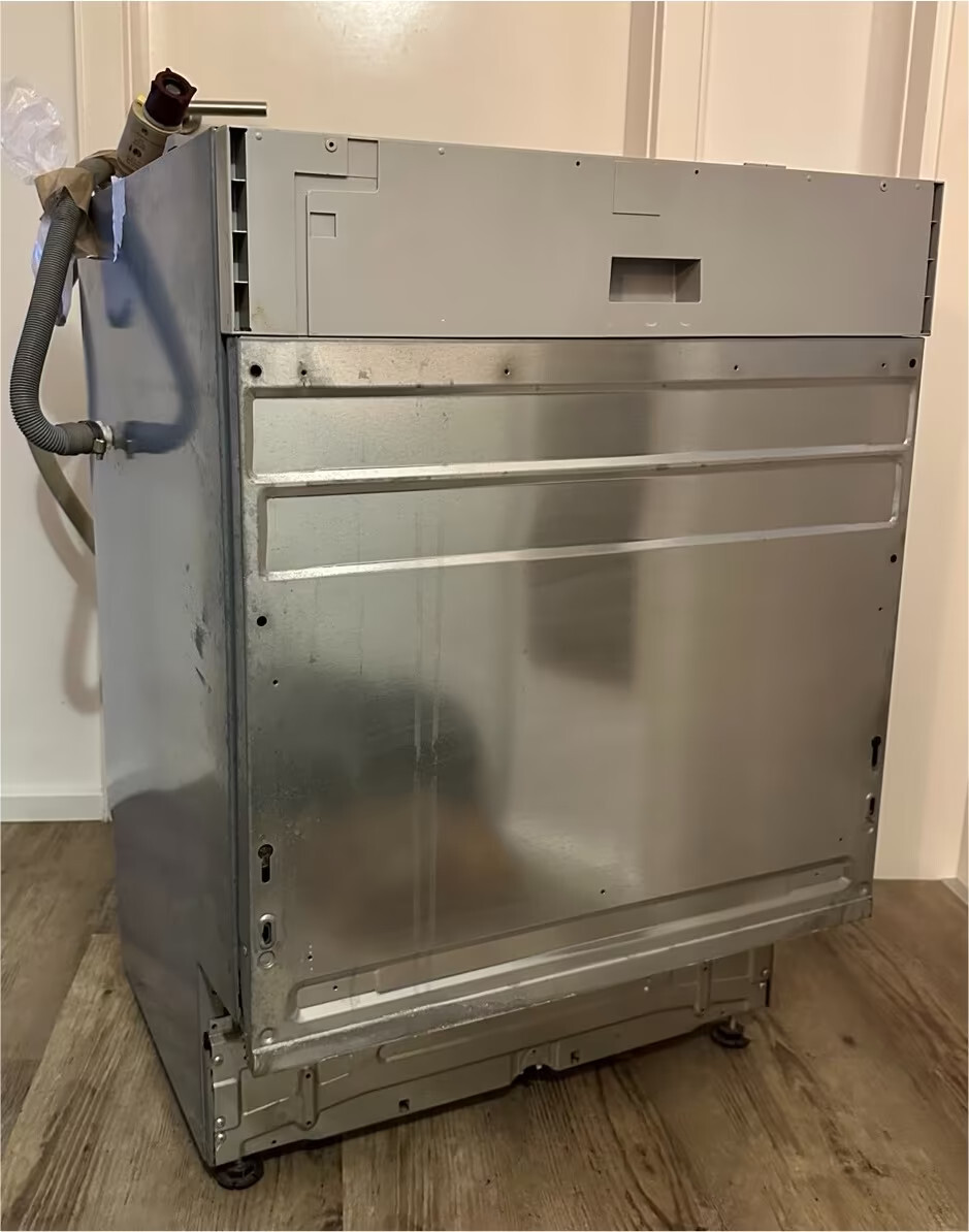 Metallische Spülmaschine auf Holzboden mit Schlauchanschluss links.
