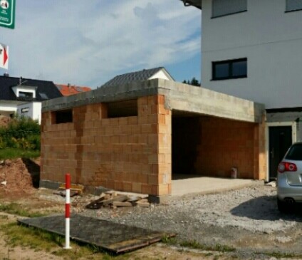 Kleine offene Garage im Rohbau aus Ziegelstein mit Betonboden neben Wohnhaus und Auto