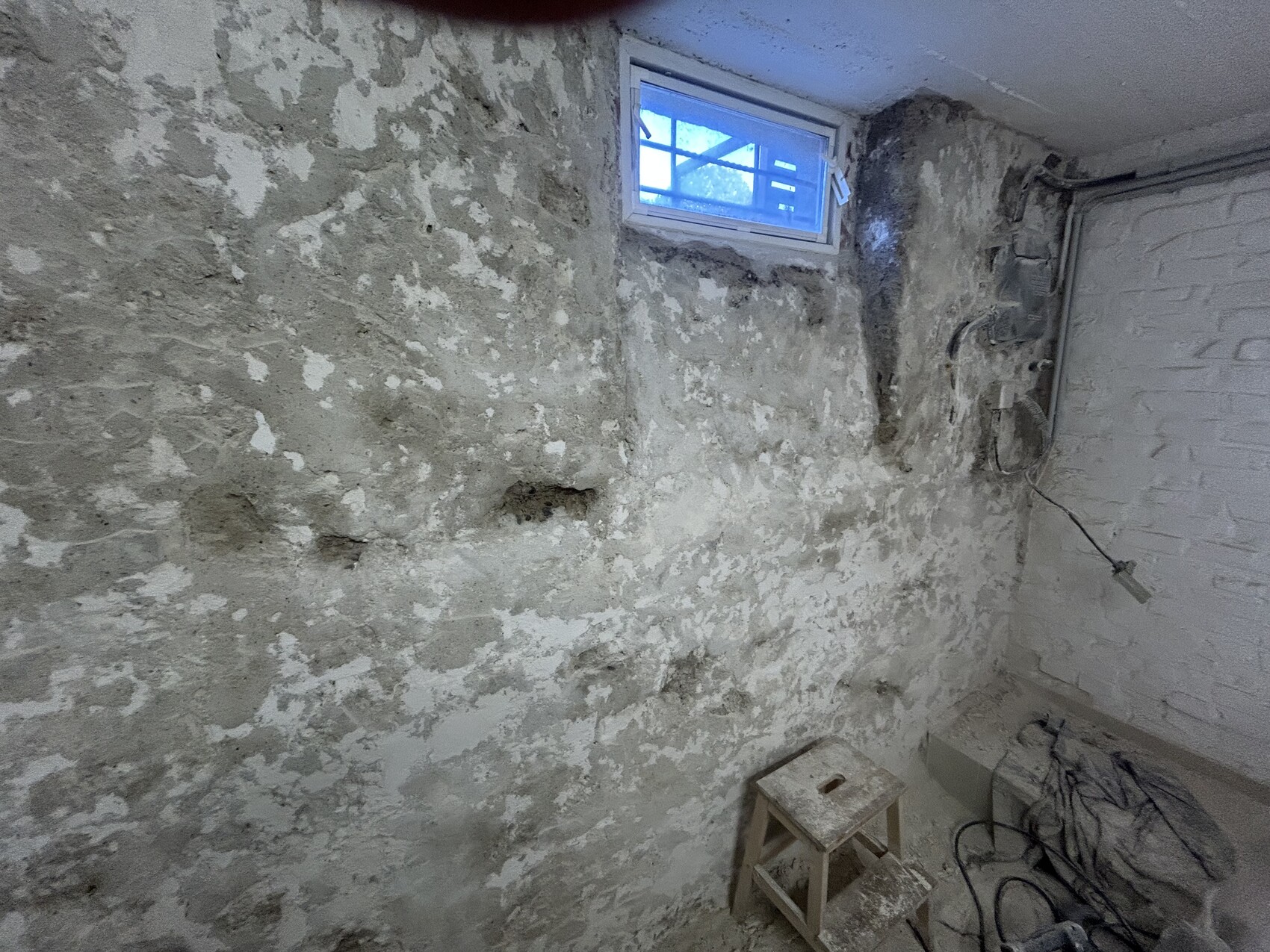 Basement room with peeling plaster on rough wall, window and cables visible