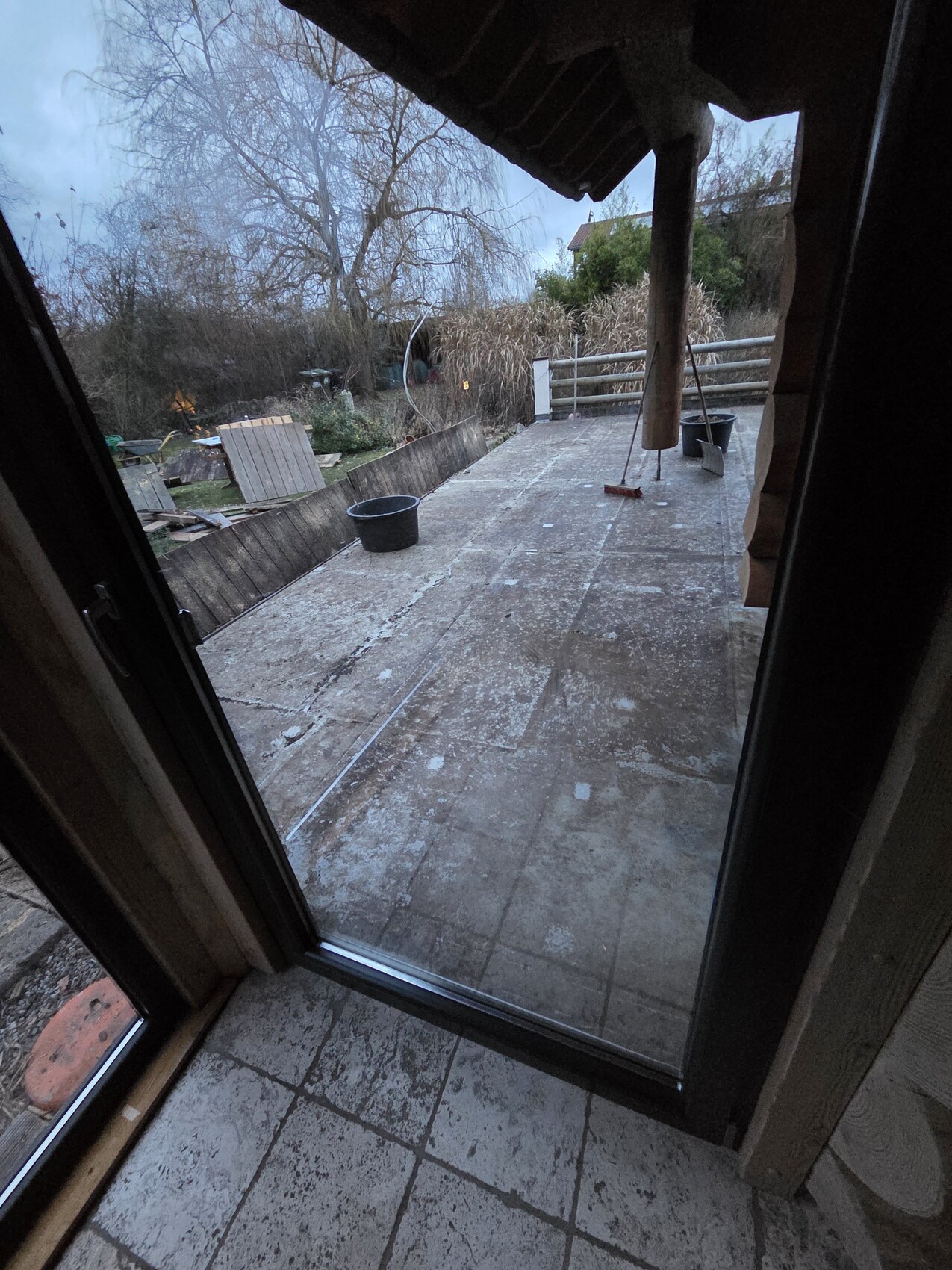 Blick aus dem Innenraum auf eine verwitterte Terrasse mit Eimern, Besen und Geländer im Hintergrund