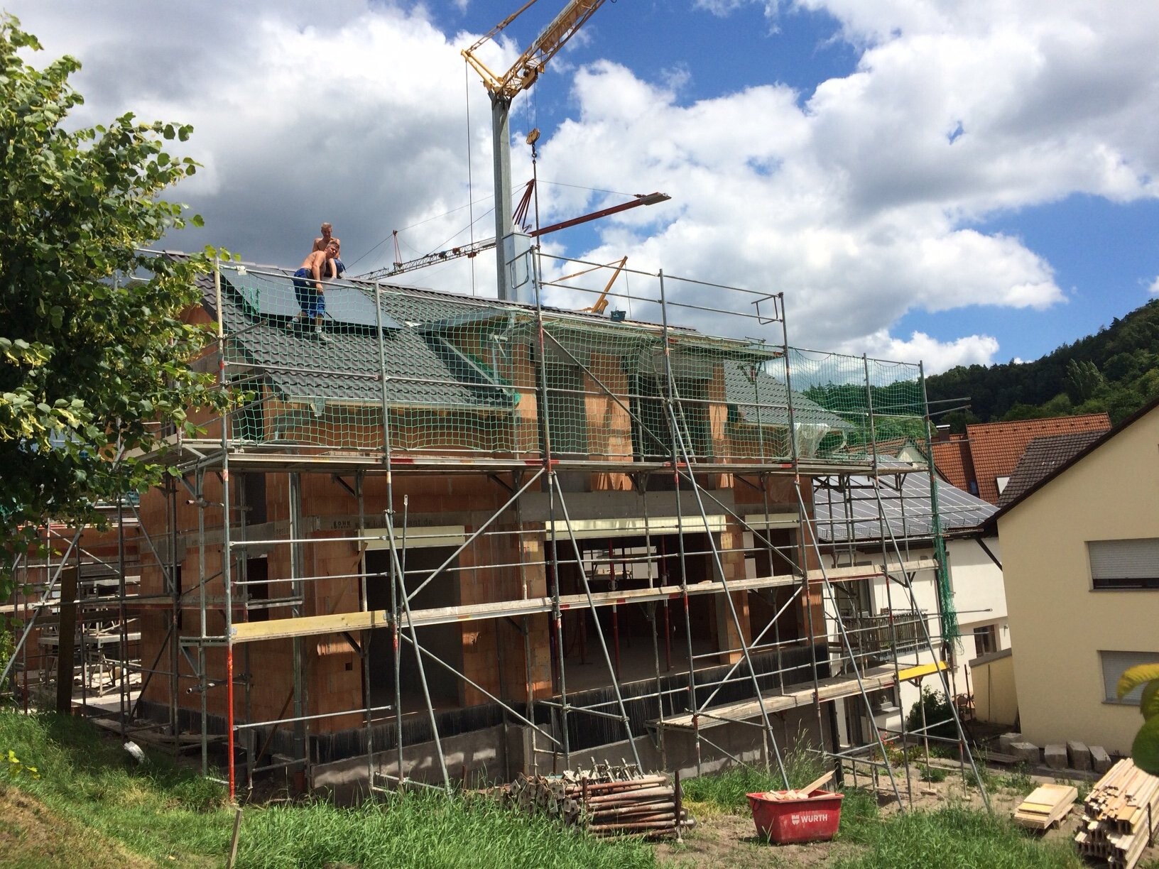 Construction site with scaffolding, two workers on the roof, crane in the background, surrounding houses