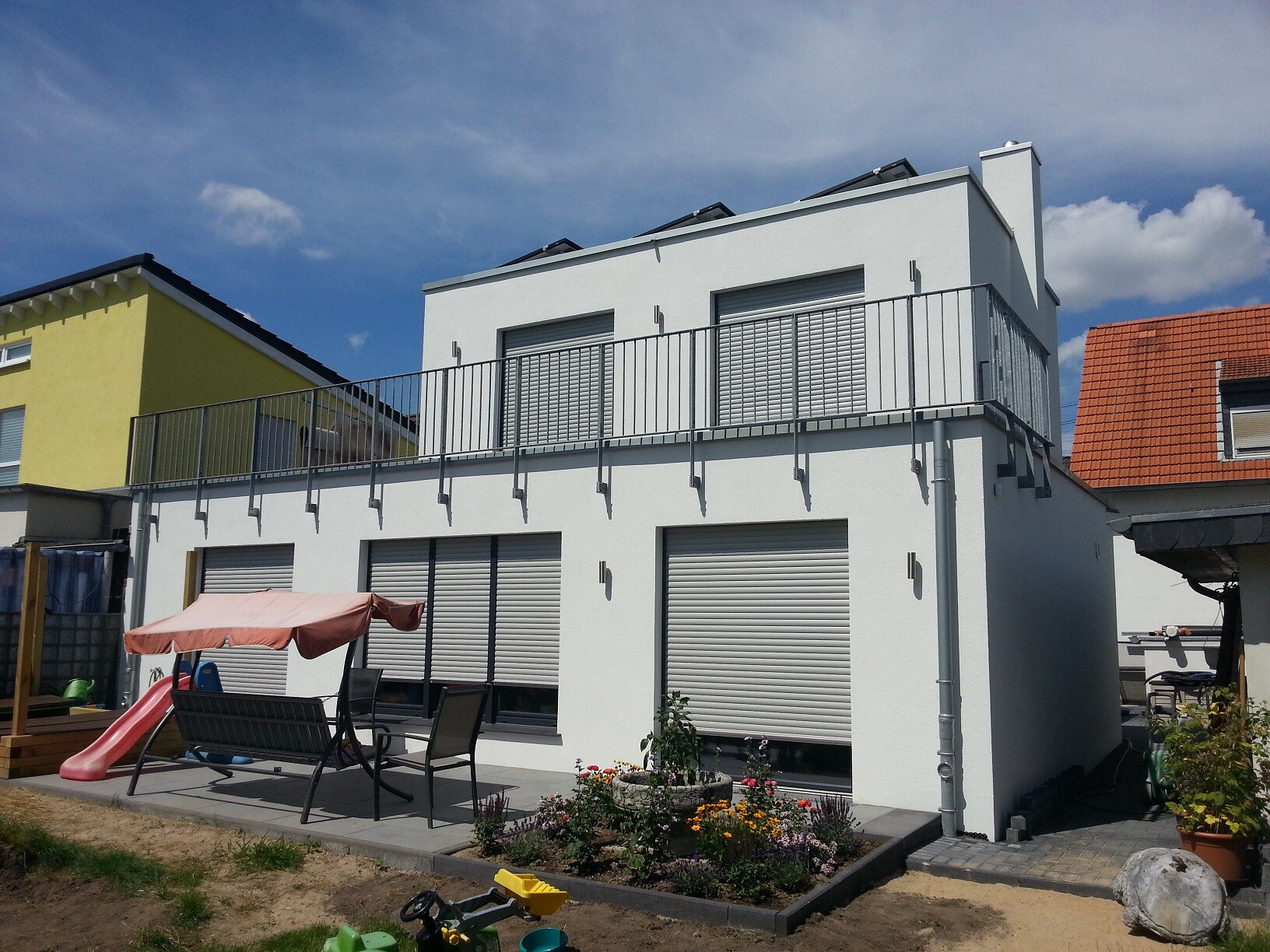 Modern white villa with balcony, terrace, garden furniture, play equipment, and flower bed.